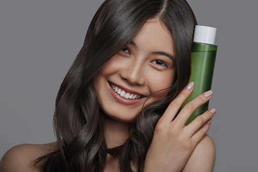 Close up portrait of beautiful young asian woman with clean fresh skin holding green cosmetic tube near her face on gray background. Face care, Facial treatment, Cosmetology, beauty and spa, Asian women portrait.