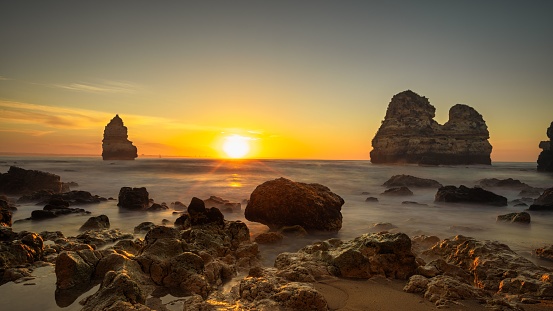 A stunning shot of Camilo Beach in Algarve, Portugal featuring a rocky shoreline illuminated by a vivid orange sunset