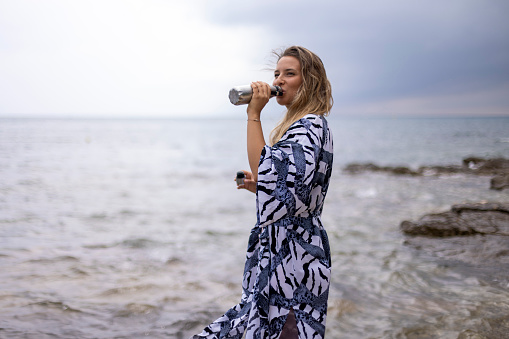 Cheerful woman drinks water from a reusable bottle while standing on the seashore