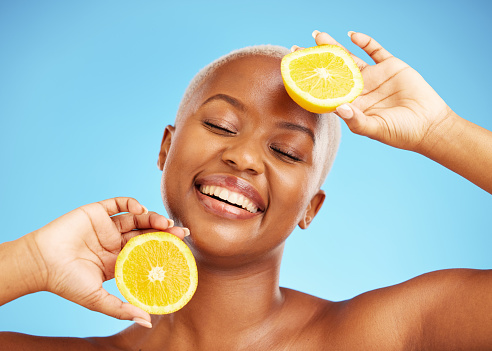 Orange, face and nutrition with a model black woman in studio on a blue background for treatment. Beauty, skincare and smile with a happy young person holding natural fruit for health or wellness