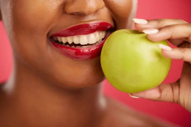 Photo of Woman, mouth and apple for natural nutrition, diet or health and wellness against a red studio background. Closeup of female person smile with lipstick and organic fruit for vitamin, fiber or food