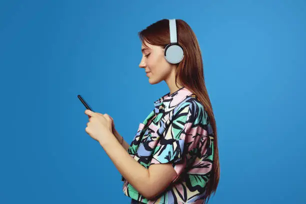 Photo of Side view of young student girl holding mobile phone and wearing headphones while listening to music