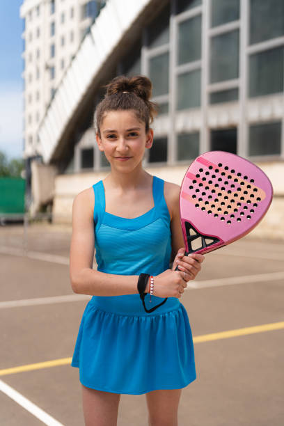Happy young girl standing with padel racket on sports ground near building stock photo
