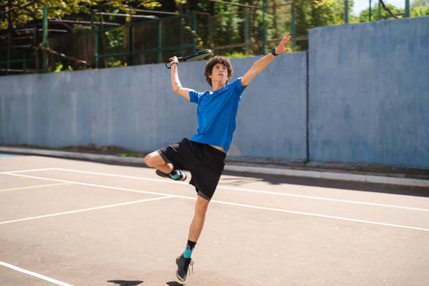 Teenage boy jumping on tennis court and hitting with racket on summer day stock photo