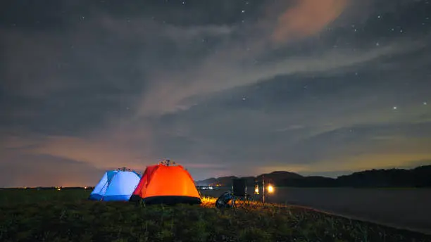 Photo of riverside camping, blue and orange riverside tent camping with luggage, bonfire, evening camping activities The atmosphere of camping at night when the sky is full of stars.