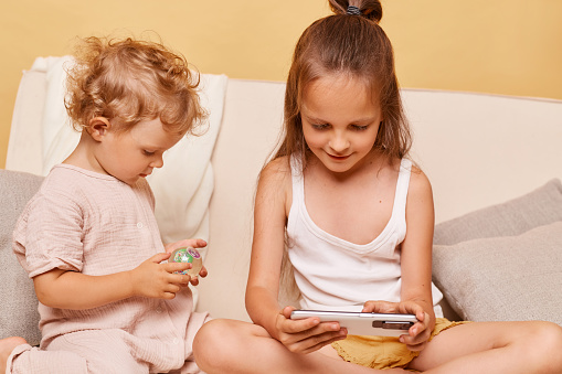 Closeup portrait of charming curious little girls sisters using phone while sitting on sofa at home against beige wall using smartphone browsing internet kids content.
