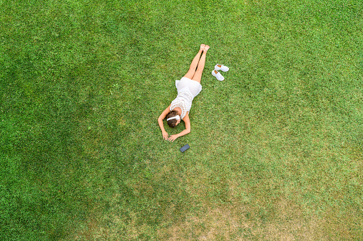 Woman in a white dress and headphones, lying on the green grass sunbathes at summer day. Top view, drone, aerial view
