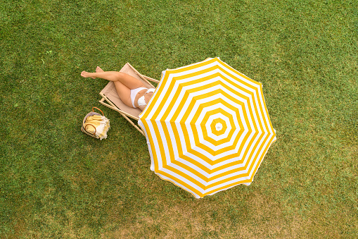 Woman in a white bikini with basket for picnic sitting on deck chair under yellow umbrella  on the green grass sunbathes at summer day. Top view, drone, aerial view.