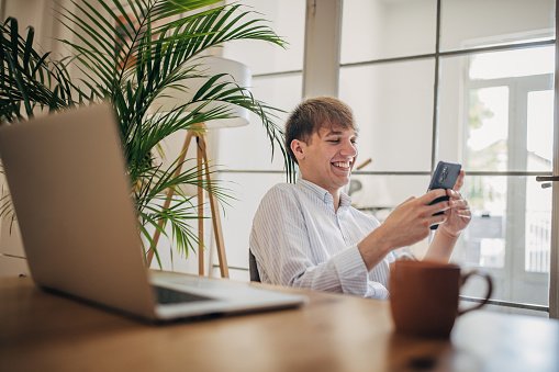 Modern young man using smart phone in home office while sitting at the desk.