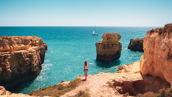 Woman standing on rock enjoying beautiful view of atlantic ocean, rock formation and yacht- travel,  summer vacation in Portugal