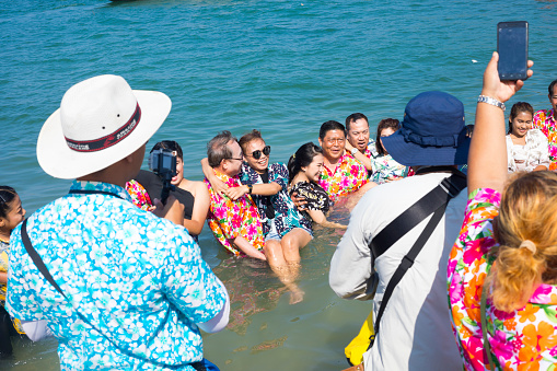Some thai people are photographing happy group  of thai men standing and carrying women in sea at beach of small neighbor island to Ko Sichang. Scene is during local Songran festival and people are fully dressed. Men are carrying women for putting them slowly into water as form of traditional local style as island is fisher men island and women symbolize meermaids