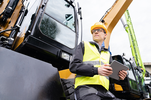 Engineer in a helmet with a digital tablet next to construction excavators