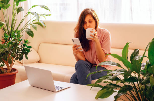 istock Young woman using smartphone and laptop, drinking coffee, sitting on sofa between green plants in the morning. 1578547769