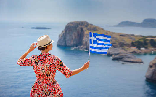 Nice Happy Female Enjoying Sunny Day on Greek Islands. Travel to Greece, Mediterranean islands outside tourist season. Young traveling woman with national greek flag enjoying view on sea