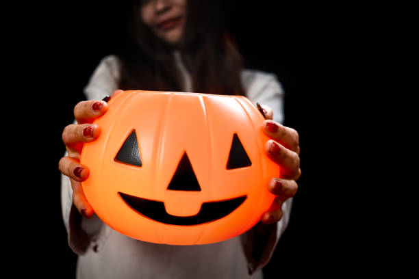Halloween orange pumpkin in hand of beautiful attractive black long hair woman in spooky white ghost costume standing in dark room, female with makeup scary faces going to celebrate Halloween party. calabaza naranja de halloween en la mano de la hermosa y atractiva mujer negra de cabello largo con espeluznante disfraz de fantasma blanco de pie en la habitación oscura, mujer con caras de miedo maquilladas que van a celebrar la fiesta de halloween.