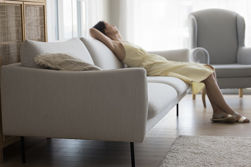 Relaxed young woman, teenage girl resting on soft comfortable sofa, enjoying leisure in stylish living room with new furniture of pale colors, stretching body, breathing fresh air