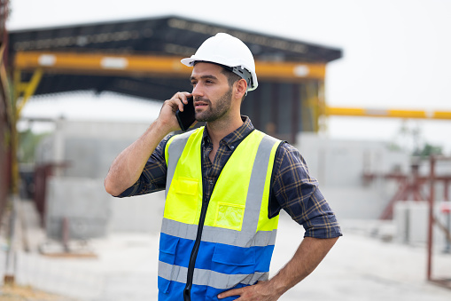 building and construction worker using mobile phone, Hiapanic latin male wearing safety hard hat helmet standing with arms crossed at construction site and looking at camera