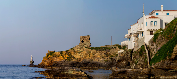 Seascape. View of famous, urban, ancient lighthouse in the city Chora, sea and fortress on a sunny spring evening (Greece, island Andros, Cyclades)