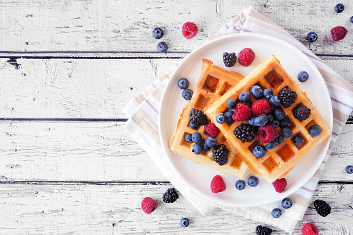 Breakfast waffles with raspberries and blueberries. Overhead view table scene on a rustic white wood background.