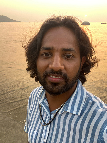 Stock photo showing an Indian man on holiday in Goa, South India, pictured standing on Palolem Beach, paddling in the gentle sea waves at sunset, a particularly popular winter holiday destination for both English and German tourists.