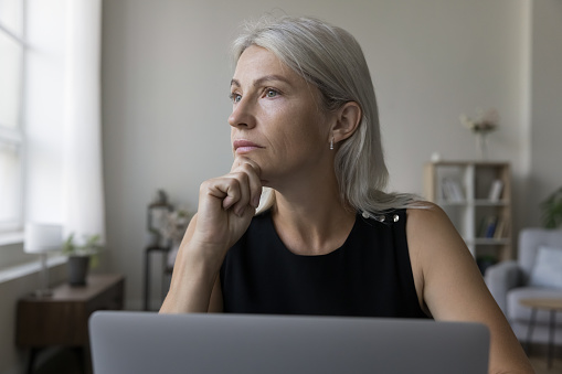 Serious middle aged freelance business woman working at laptop at home, thinking over creative ideas for project, looking away, touching chin, using gadget for job Internet communication