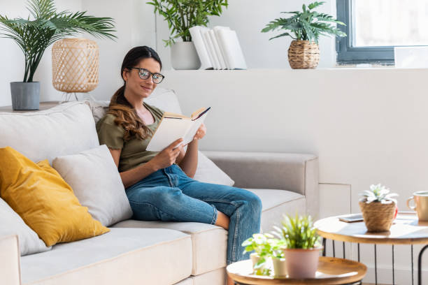istock Confident woman reading a book while sitting on sofa at home. 1578914084