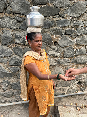 Stock photo showing an irregular stone wall in front of which an Indian woman wearing Salwar Kameez traditional clothing is stood with a metal pot of water on her head.