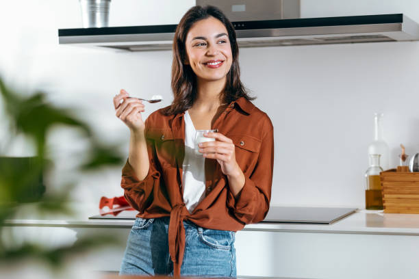Beautiful happy woman eating a yoghurt while looking forwards in the kitchen at home Shot of beautiful happy woman eating a yoghurt while looking forwards in the kitchen at home yogurt stock pictures, royalty-free photos & images