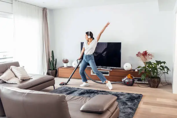 Photo of Young happy woman listening and dancing to music while cleaning the living room floor with a vaccum cleaner