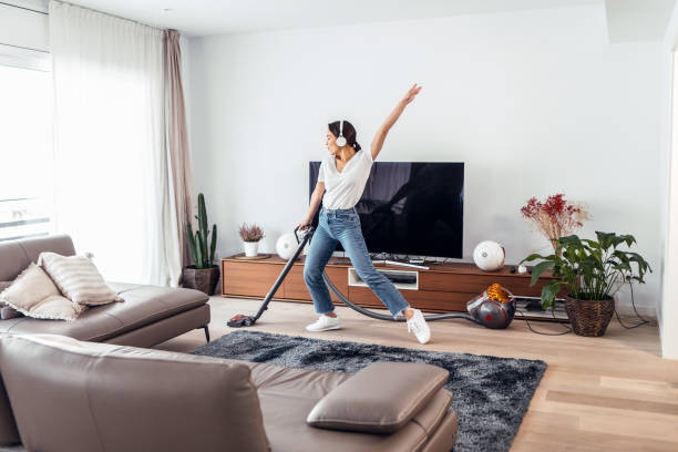 young happy woman listening and dancing to music while cleaning the living room floor with a vaccum cleaner - spring cleaning women cleaning dancing imagens e fotografias de stock