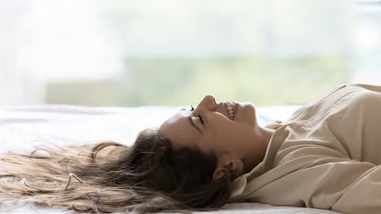 Happy young woman resting on comfortable mattress in bed, lying on back, smiling, laughing with closed eyes, enjoying relaxation, leisure, weekend, vacation in hotel suite
