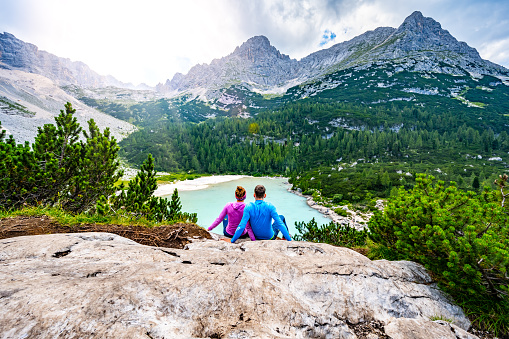 Description: Young sportive couple enjoys view on the turquoise Sorapis lake from a beautiful restpoint in the afternoon. Lake Sorapis, Dolomites, Belluno, Italy, Europe.