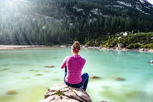 Description: Woman sits on big rock and enjoys the view at beautiful turquoise Sorapis lake in the afternoon. Lake Sorapis, Dolomites, Belluno, Italy, Europe.