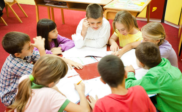 Group of elementary school children sitting around the table in the classroom and reading. 