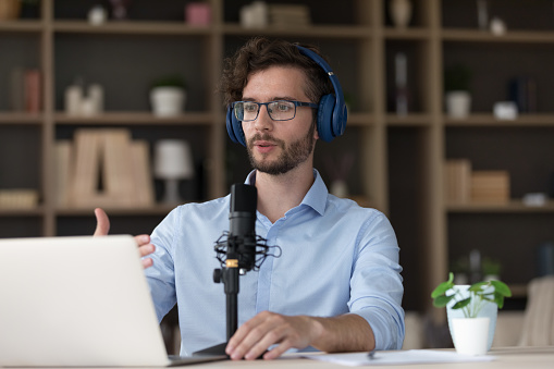 Serious handsome young businessman using wireless headphones, sound studio big microphone, laptop for video call, speaking on online conference. Professional influencer broadcasting online