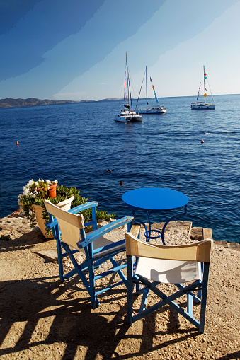 Idyllic  view on Free table in the cafe and chairs  in colors of Greece - white and blue. People swimming in distance  and yachts (sailboats) anchored behind
