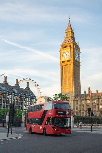 London, England - June 23, 2023: Red double-decked bus on the road in front of the Big Ben tower and London Eye ferris wheel at sunset