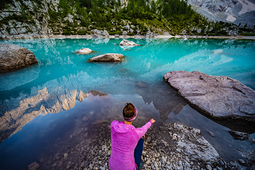 Description: Young woman enjoys beautiful refelctions on turquoise Sorapis lake and dito di dio in the evening. Lake Sorapis, Dolomites, Belluno, Italy, Europe.
