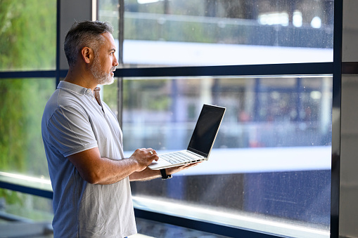 Mature businessman with blank expression working on a laptop while standing at a window