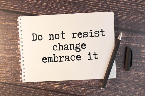 a white notebook with the text Do Not Resist Change Embrace It lies on a wooden surface near a white keyboard with a red marker.