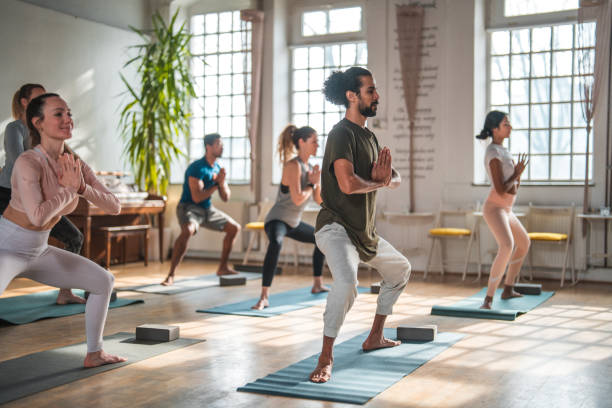 Fashionably Dressed Biracial Man With a Group of Mid Adult Sporty People in Yoga Class Mixed group of sporty,  multiracial mid adult men and women working out together in a modern gym. 3/4 length image. Side view. Exercise  stock pictures, royalty-free photos & images