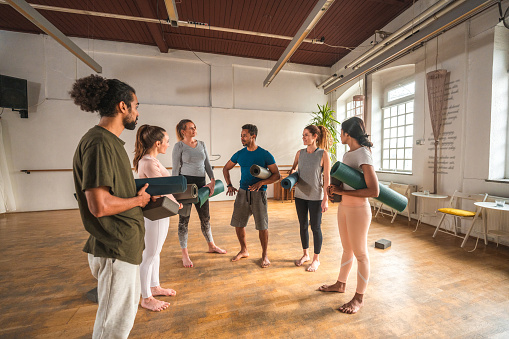 Attractive multiracial group of men and women in sports wear standing in a circle, talking to each other at yoga gym. 3/4 length image.