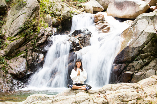 A mid age Caucasian woman in zen like meditation on rocks in front of a waterfall.