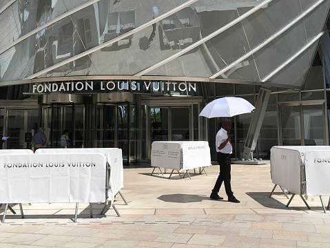 Paris, France - July 8, 2019 : Employee of the Fondation Louis Vuitton shielding himself from the sun with a white umbrella in front of the museum's entrance on a hot summer day in Paris, France