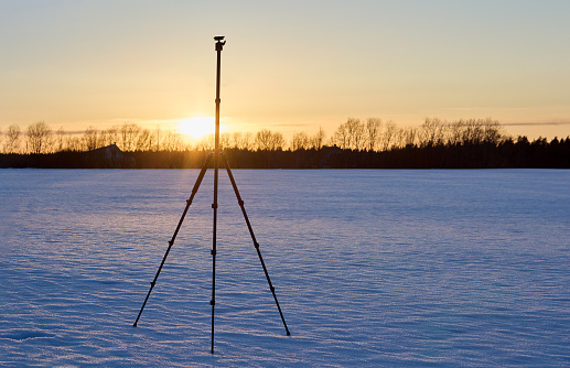 Photographic tripod is set outdoors in winter and stands on snowy crust in field.