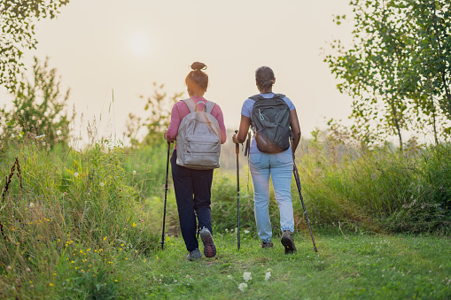 A mother and her daughter take a hike on a warm summers evening as they stay fit together.  They are both dressed casually, are carrying backpacks and have walking sticks in hand.