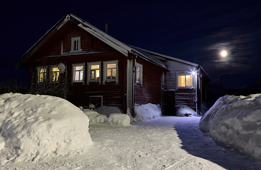 Snow covered country house with electric light in windows on moonlit night.