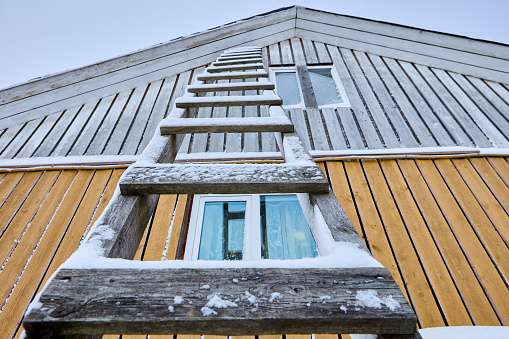 Wooden scaling ladder with steps is leaning against wall of wooden country house in winter.