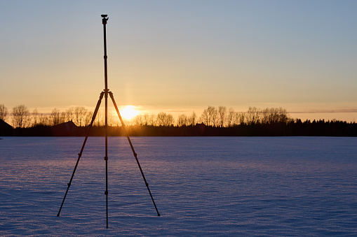 Photographer equipment, an aluminum tripod for camera is set on snowy crust in field after sunset.