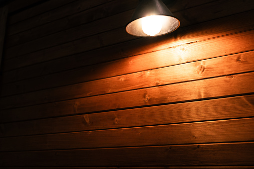 There is a wall lamp on the wooden wall, and the yellow light illuminates a part of the wooden board.